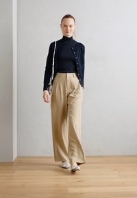 Un mannequin porte un col roulé noir, un cardigan bleu marine et un pantalon large beige, assortis à des ballerines argentées et un petit sac à bandoulière.