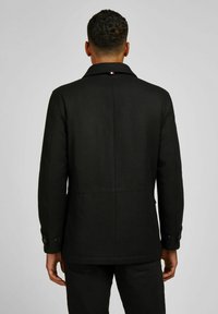 Man gezien vanaf de achterkant, gekleed in een zwarte jas en zwarte broek, staand tegen een effen lichtgrijze achtergrond.