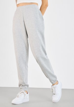 Personne portant un pantalon de jogging gris clair à taille élastique et des baskets blanches, debout avec une jambe légèrement pliée et les mains dans les poches.