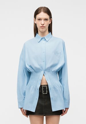 Woman wearing a light blue button-up shirt with a cinched waist, paired with a black mini skirt and a wide belt with a rectangular buckle.