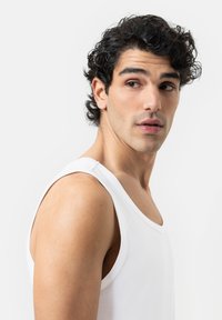 White cotton tank top with wide straps, smooth texture, and a round neckline worn by a model facing slightly sideways against a plain background.