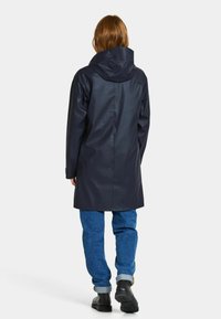 Marinblå vattentät regnjacka med huva, slät textur, mittlång design och sidofickor, i kombination med blå denimbyxor och svarta skor.