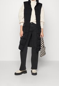 Czarna długa kamizelka puchowa z wysokim kołnierzem, zestawiona z kremowym swetrem i czarnymi dżinsami. Do tego kraciasta torba i masywne beżowe buty.