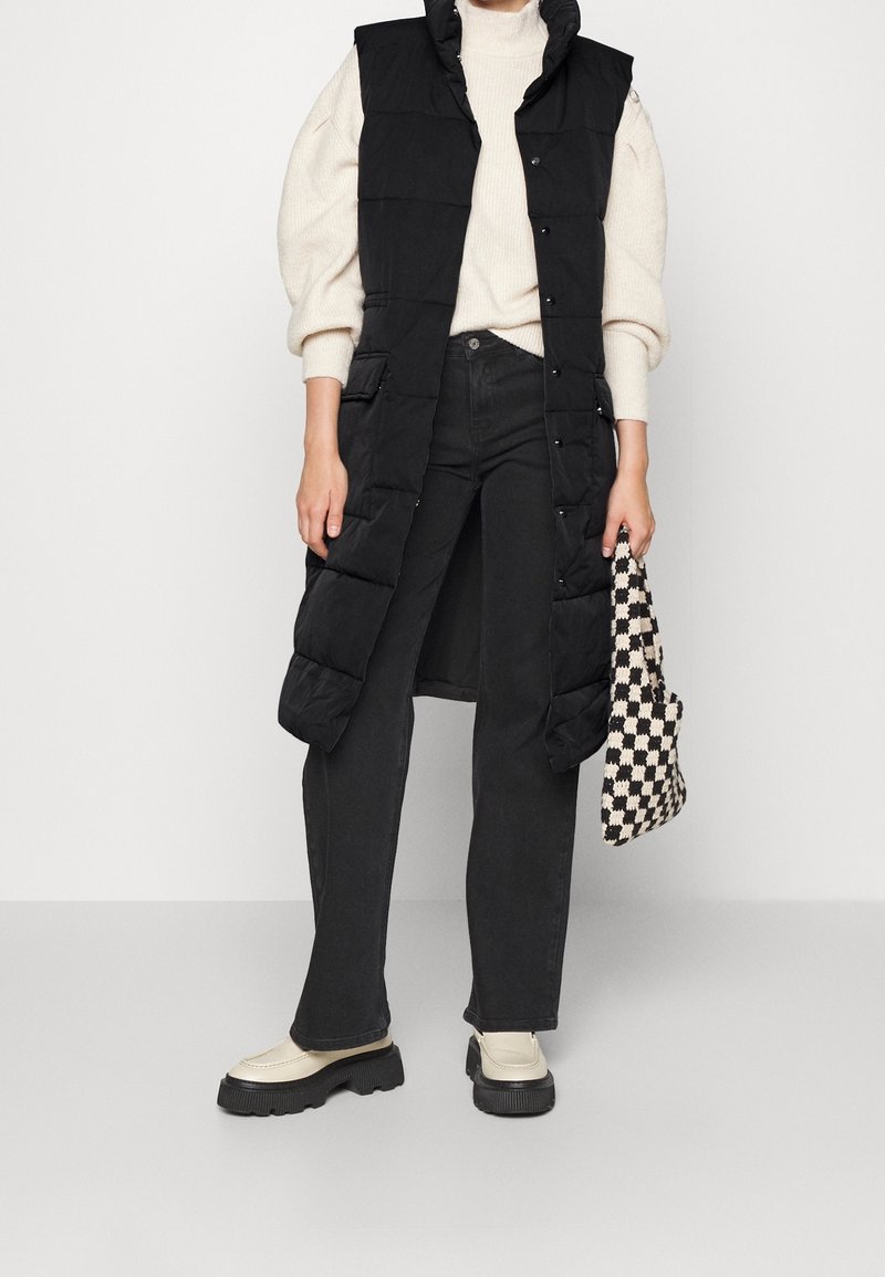 Czarna długa kamizelka puchowa z wysokim kołnierzem, zestawiona z kremowym swetrem i czarnymi dżinsami. Do tego kraciasta torba i masywne beżowe buty.