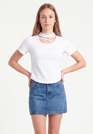 Jeune femme aux longs cheveux portant un haut blanc à manches courtes avec des découpes et une jupe en jean bleue, debout les mains sur les hanches sur fond blanc.