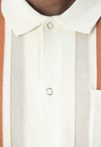 Polo de punto con base crema, que presenta rayas verticales en beige y naranja, un cuello puntiagudo y un bolsillo en el pecho con un botón metálico.