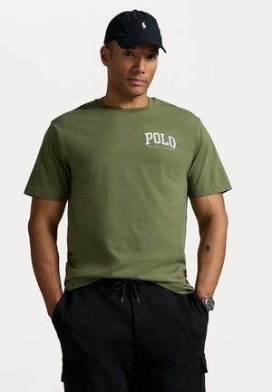 Olive zeleno bombažno majico s belim logotipom "Polo Ralph Lauren". Ima kratke rokave in okroglo ovratnico. Ujemajo se s črnimi hlačami in uro.