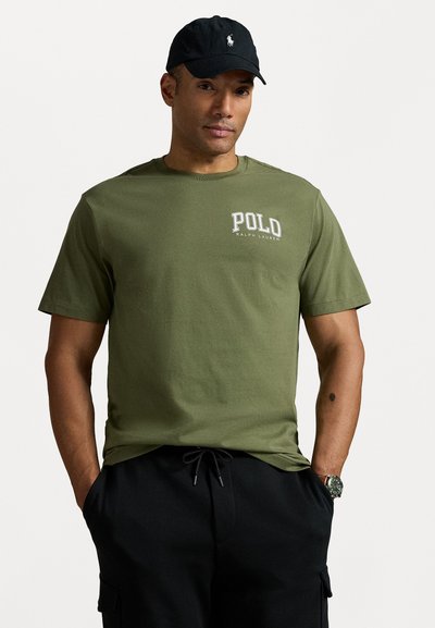 Olivenfarget bomulls T-skjorte med "Polo Ralph Lauren" logo i hvitt. Korte ermer og rund hals. Kombinert med svarte bukser og klokke.