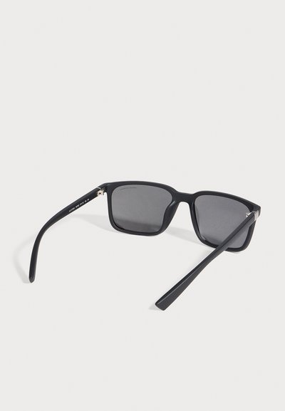 Lunettes de soleil noires rectangulaires, aux verres gris foncé et aux branches ouvertes, sur un fond blanc uni.