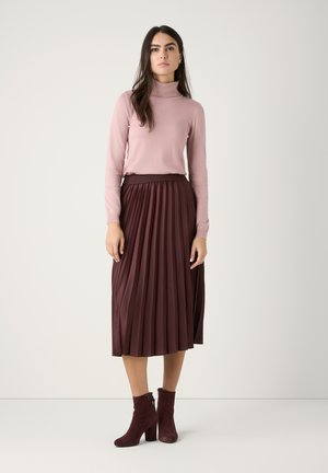 Un pull à col roulé en tricot rose clair associé à une jupe midi plissée bordeaux. Le mannequin porte des bottines à talons en suède bordeaux foncé.