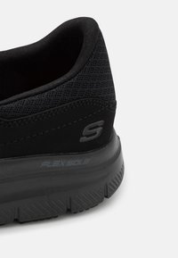 Zapatilla deportiva negra con parte superior de malla transpirable, suela flexible y suela de goma texturizada. Presenta un logo y un talón reforzado para mayor soporte.