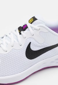Scarpa da ginnastica bianca con tomaia in mesh, logo swoosh nero, accento viola sulla suola e lacci a contrasto con un'etichetta nera e gialla.