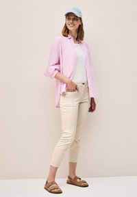 Camisa de rayas rosa claro sobre una camiseta blanca, combinada con pantalones color crema y sandalias marrones. Se lleva una gorra azul clara en la cabeza.