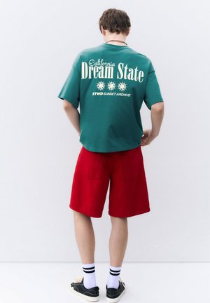 Ung mand set bagfra iført en grøn "California Dream State" t-shirt, røde shorts, hvide sokker og sorte sneakers mod en ensfarvet baggrund.