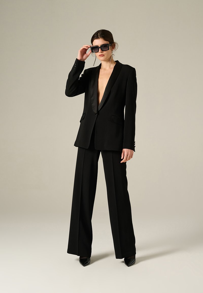 Completo nero con blazer a profondo scollo a V e pantaloni a gamba larga. Caratterizzato da rever in satin e tasche sottili. Modello che indossa occhiali da sole oversize.