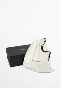 Boîte rectangulaire noire et pochette blanche avec cordons noirs, toutes deux étiquetées "Massimo Dutti", sur fond blanc.