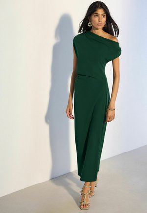 Žena v tmavozelenej midi šatách na jedno rameno s béžovými sandálmi na podpätku stojaca pri svetlej stene so tieňom za ňou.