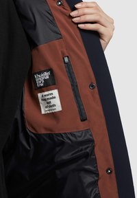 De binnenkant van de zwarte en bruine jas heeft een gladde voering, gelabelde patches, een ritszak en een stoffen label met zorginstructies.