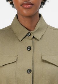 Ελιά πράσινη μπλούζα με αιχμηρό γιακά, δύο μπροστινές τσέπες και μαύρα κουμπιά με κυκλικό σχέδιο. Υφή που μοιάζει με βαμβάκι.