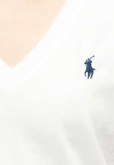 Close-up af en person iført en hvid V-hals skjorte med et lille broderet marineblåt polospillerlogo på brystet.