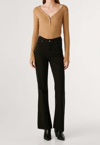 Femme portant un haut beige à manches longues avec un col en V, un pantalon évasé noir taille haute et des bottes noires à bouts pointus, debout sur un fond blanc.