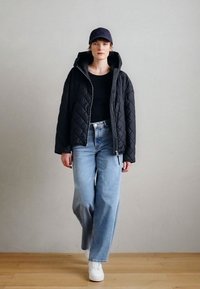 Čierna prešívaná bunda so zipsom vpredu, v kombinácii s priliehavou čiernou košeľou a svetlomodrými džínsami s širokými nohavicami. Celý outfit dopĺňajú biele tenisky.