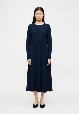 Ženska stoji obrnjena naprej, oblečena v mornarsko modro midi obleko z dolgimi rokavi in črnimi čevlji s koničastimi konicami na belem ozadju.