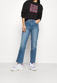 Felpa nera con scritta colorata "FEELIN' DARN GOOD", abbinata a jeans denim a zampa chiari e sneakers bianche con dettagli rossi e blu.