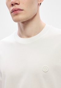 Tricou alb cu mâneci scurte, realizat dintr-un material moale, având un decolteu rotund și un logo mic în relief pe piept.