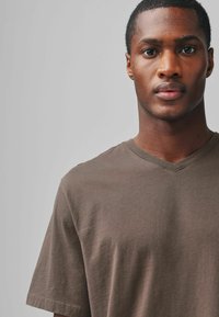Tricou din bumbac maro cu decolteu în V, mâneci scurte și o textură netedă, prezentat pe un fundal neutru.