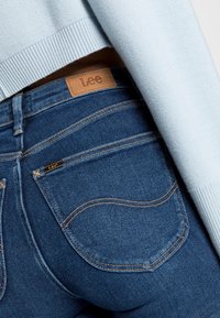 Jeans ajustados de mezclilla azul con un parche de cuero marrón de la marca Lee en la cintura, que presentan costuras en contraste y diseños de bolsillos traseros curvados.