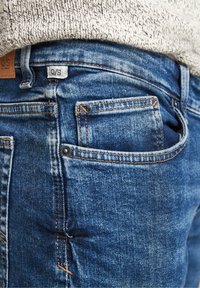 Blue denim jeans feature a back pocket with a small button detail and contrasting orange stitching, labeled with a "Q/S" tag.