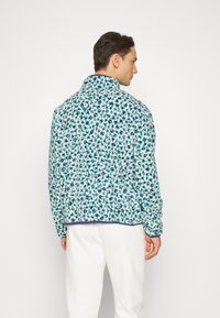 Jachetă matlasată cu un imprimeu leopard albastru și verde, având un guler înalt și o margine contrastantă la tiv. Purtați-o peste pantaloni albi.