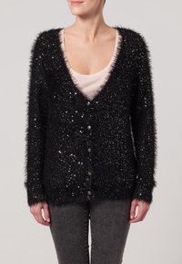 Svart fluffig cardigan med paljetter, v-ringad design, knappstängning, långa ärmar och textilstruktur. Bärs över en beige topp.