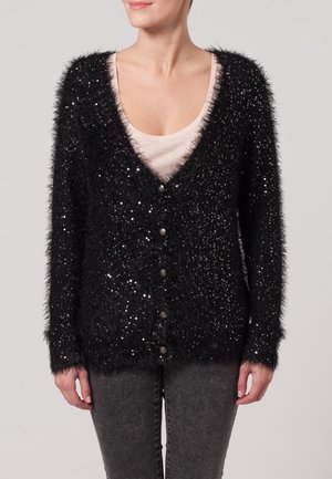 Cardigan nero peloso con paillettes, design a scollo a V, chiusura con bottoni, maniche lunghe e tessuto testurizzato. Indossato sopra un top beige.