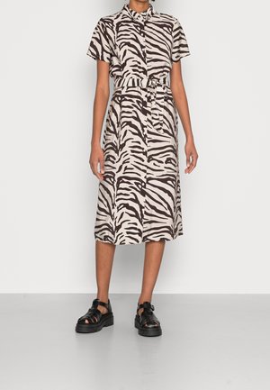Kobieta ubrana w marszczoną sukienkę z nadrukiem w paski zebry, sięgającą do kolan, z paskiem w talii i czarnymi masywnymi sandałami na paski.