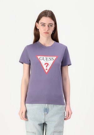 T-shirt violet à manches courtes, col rond, avec un motif de logo triangulaire rouge portant le texte "GUESS U.S.A." et un point d'interrogation.