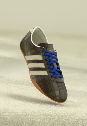 Baskets Adidas avec motif à carreaux marron, bandes blanches, lacets bleus et semelle en gomme reposant sur une surface beige texturée.
