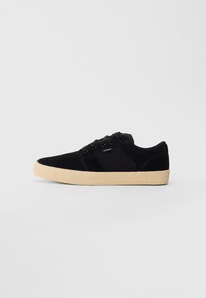 Black low-top sneaker with beige textured sole, laces, and small side logo on a plain light gray background.