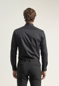 Camisa negra de manga larga, corte ajustado, textura suave y detalles de botones. Combinada con pantalones negros, vista desde la parte de atrás.
