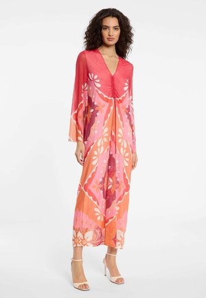 Frau steht da und trägt ein langes, durchsichtiges Kleid mit roten, pinken und orangefarbenen Blumenmustern, langen Ärmeln und beigen Sandaletten mit hohen Absätzen.