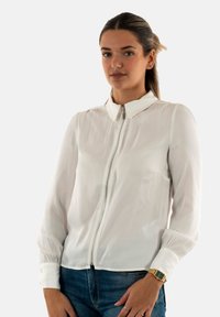 Blouse blanche à manches longues avec une fermeture éclair à l'avant, un col et des manches froncées, associée à un jean en denim foncé et une montre en or.