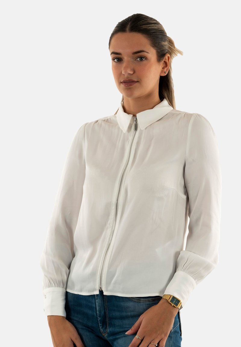 Blouse blanche à manches longues avec une fermeture éclair à l'avant, un col et des manches froncées, associée à un jean en denim foncé et une montre en or.