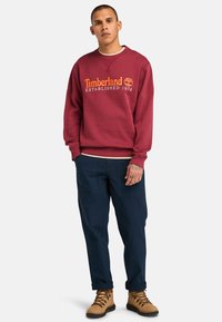 Felpa maroon con stampa arancione "Timberland Established 1973", abbinata a pantaloni blu scuro e stivali con lacci color tan.