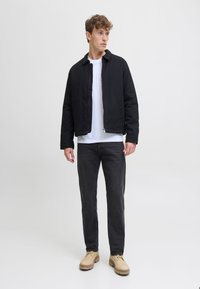 Chaqueta de algodón negra con cuello, desabrochada sobre una camiseta blanca. Tejanos gris oscuro y zapatos beige completan el atuendo. Diseño simple, sin patrones.
