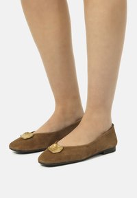 Piernas con medias nude transparentes y zapatos planos de ante marrón en punta con acentos decorativos dorados sobre un fondo blanco.