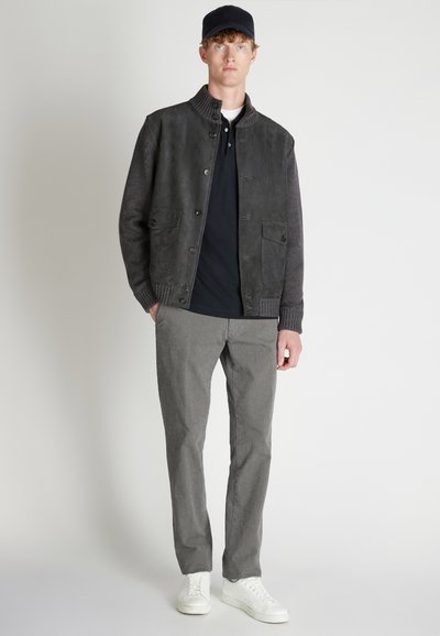 Herrenjacke aus grauem Leder und Strick mit Rippenkragen, Knopfverschluss vorne und zwei Vorderaschen. Kombiniert mit einem schwarzen Hemd und grauen Hosen.