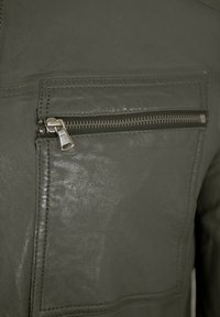 Dunkelgrüne Lederjacke mit strukturiertem Finish, einer Reißverschlusstasche und sichtbaren Steppnähten. Der Reißverschluss ist in silberfarbener Ausführung.
