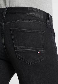 Jeans de mezclilla negra con un bolsillo trasero que tiene un pequeño parche de logo tricolor. Textura suave con un ligero desgaste en los bordes.