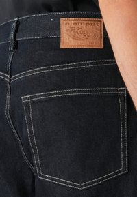Detailní záběr na zadní kapsu tmavých džínsů s kontrastním prošitím a hnědou koženou nášivkou s nápisem "element since 1992."